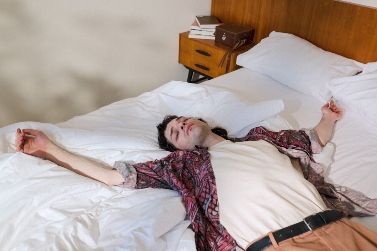 Young man laying relaxed on a bed in a vintage-style bedroom under soft daylight.