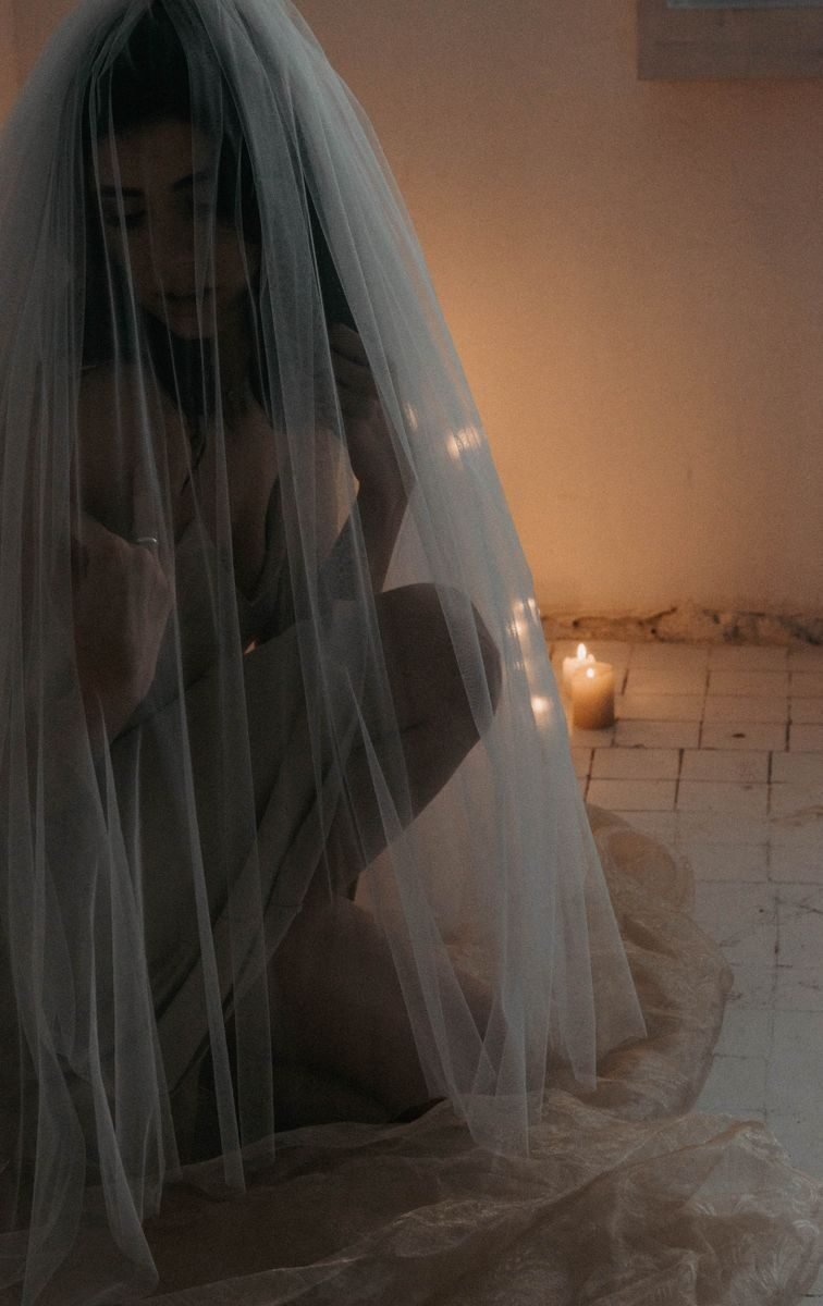 Artistic portrait of a bride kneeling under a veil, lit by candlelight creates a mysterious ambiance.