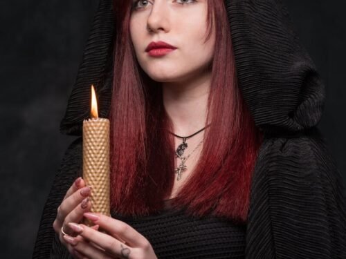 Portrait of a woman with red hair holding a lit candle, cloaked in a dark hooded garment.