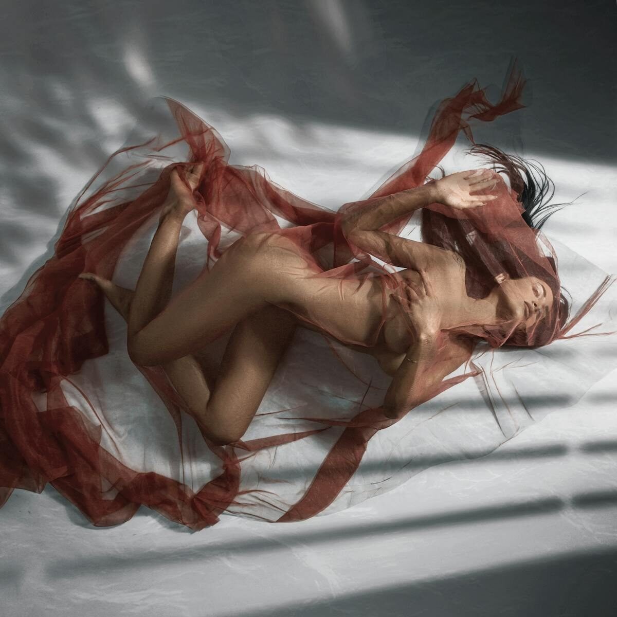 Elegant nude photography of woman with a red veil, capturing light and shadow contrast.