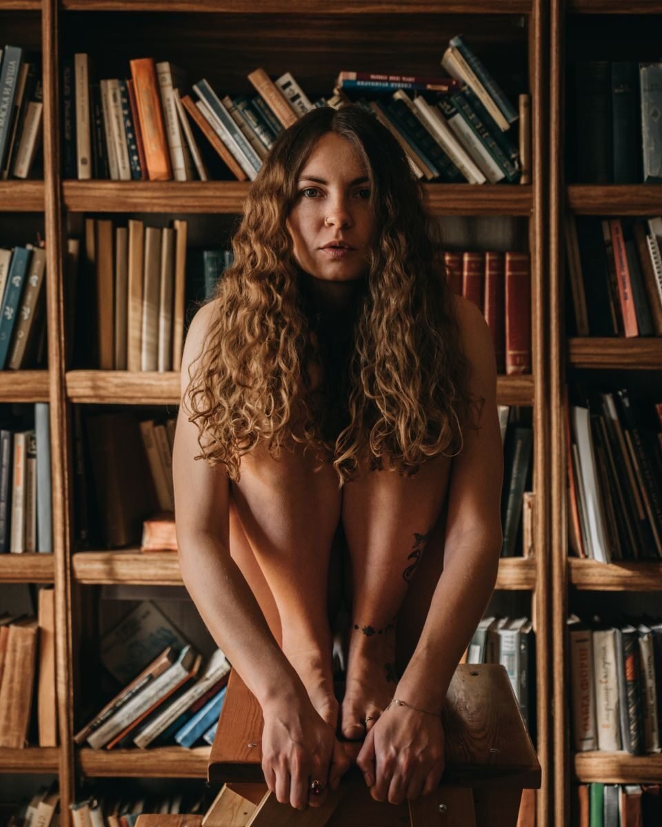 A woman with long hair sitting on a stool in a cozy library setting.