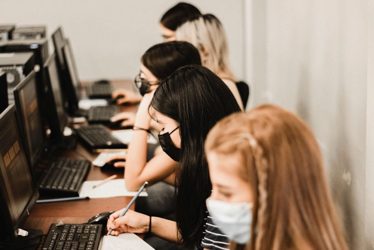 A group of students wearing face masks focus on studying in a computer lab setting.