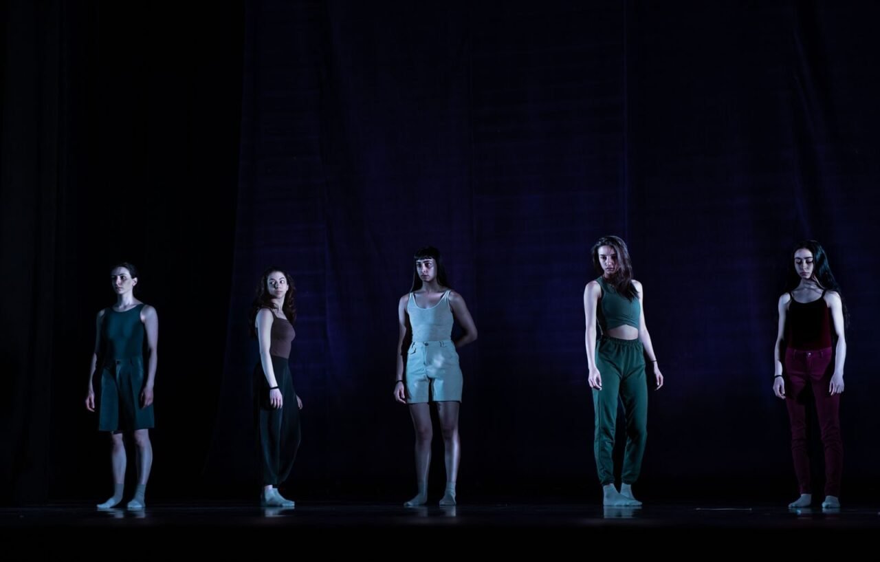 Five women dancers pose dramatically on a dimly lit stage, capturing the essence of contemporary dance.