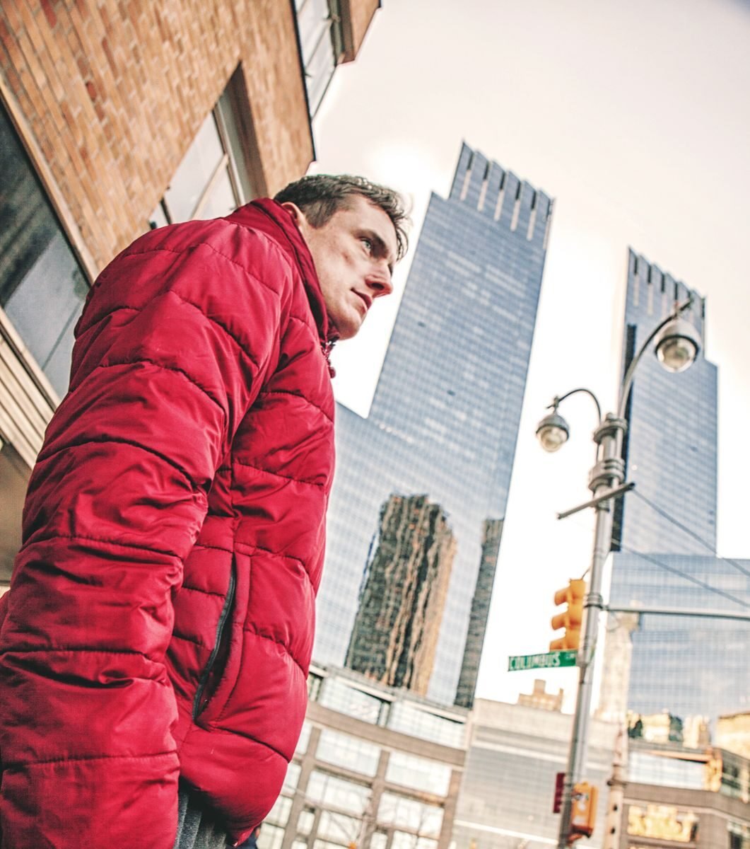 Man wearing red jacket standing against a New York City urban backdrop.