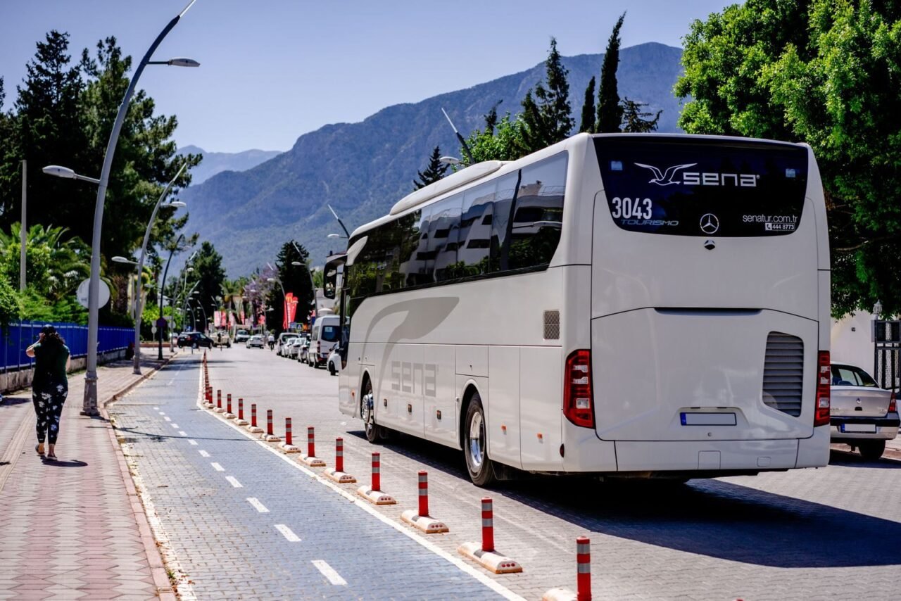 A white bus travels on a sunny mountain road lined with trees and sidewalks.