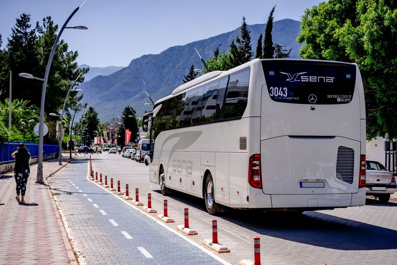 A white bus travels on a sunny mountain road lined with trees and sidewalks.