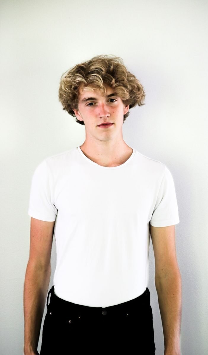 Portrait of a teenage boy with curly hair in a white shirt, standing against a white background.