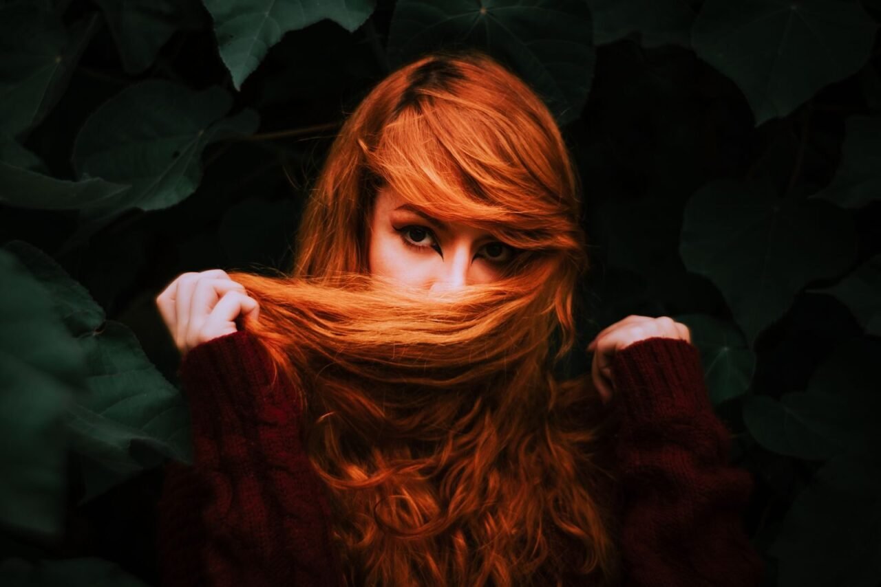 A captivating portrait of a woman with auburn hair partially covering her face, surrounded by lush green leaves.