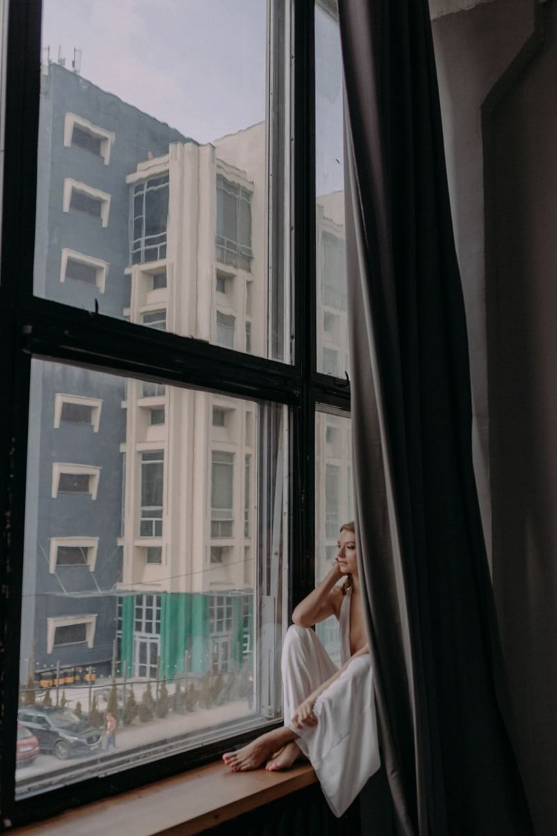 Woman in white sitting by an urban window, contemplating life, indoors.