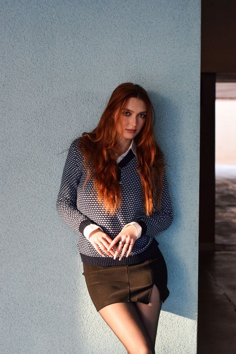 Fashionable woman with auburn hair in a chic outfit leaning on a textured wall.