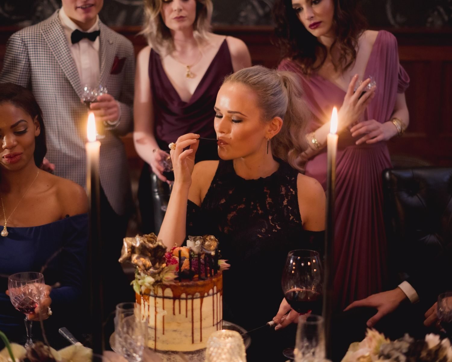 Group of elegantly dressed people enjoying a birthday celebration with cake and candlelight.