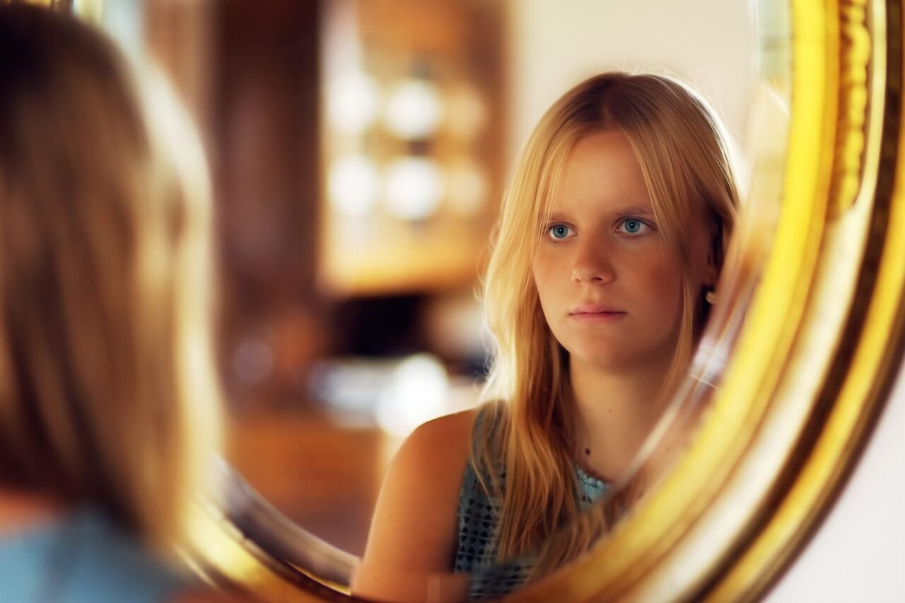 A young woman with blond hair looks at her reflection in a round mirror indoors.