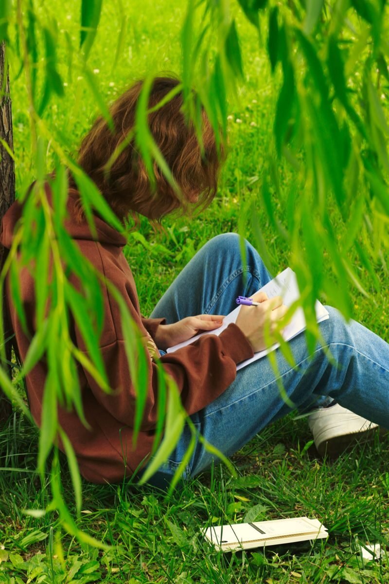 Young woman in a hoodie sits under a tree making notes on a sunny day.