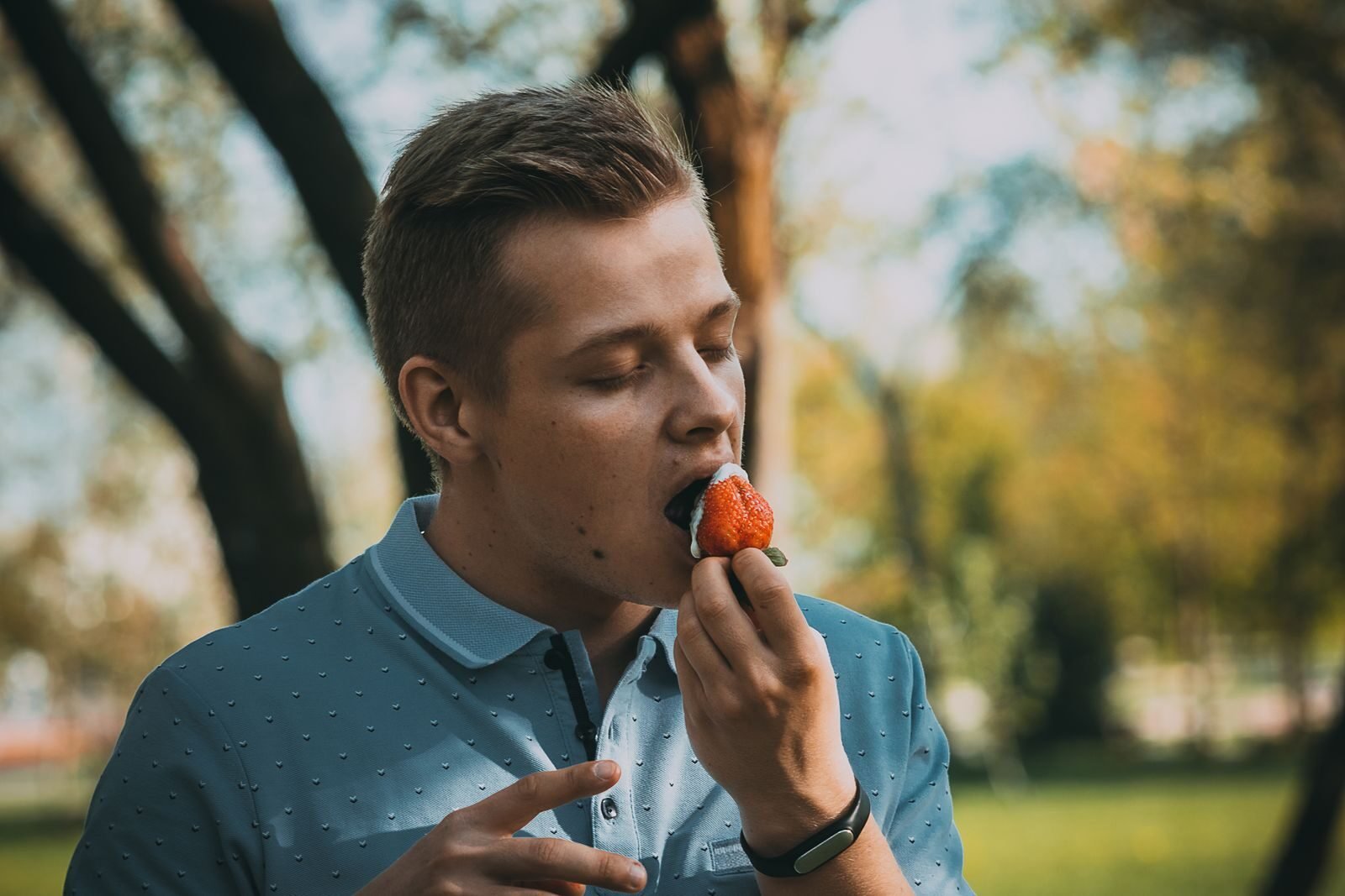 A man indulges in a ripe strawberry in a sunny park setting.