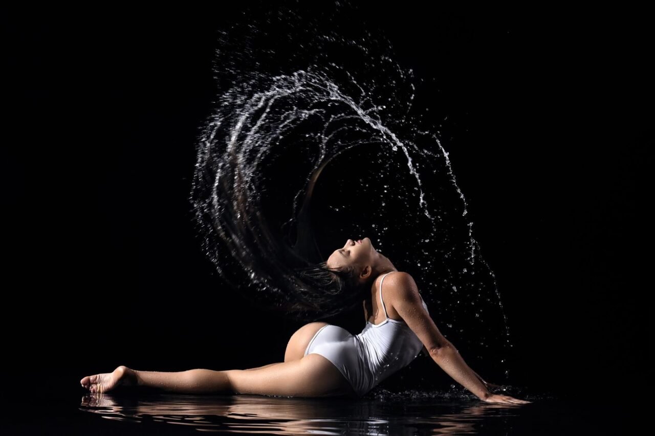 A woman in a white swimsuit poses dynamically with water splashing, against a black background.