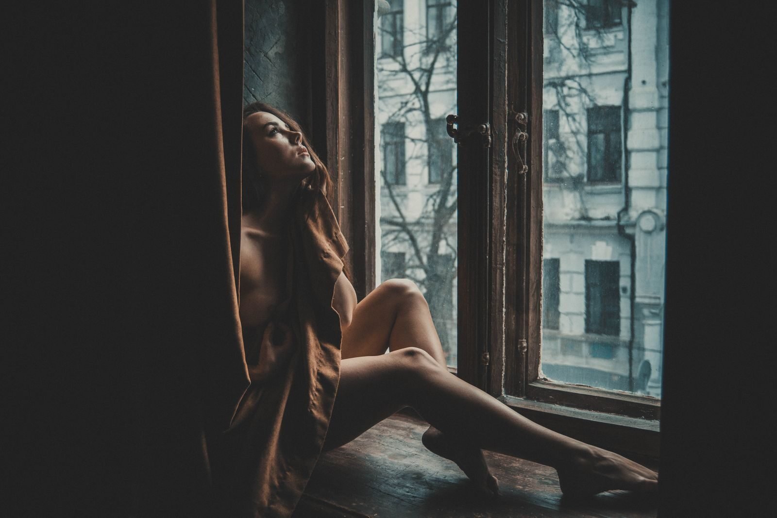 A contemplative woman sits by the window, enveloped in warm shadows, showcasing classic beauty.