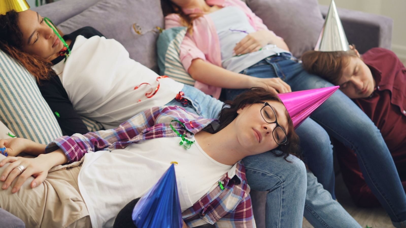 Group of young adults sleeping on a couch after a lively indoor party with hats and confetti.