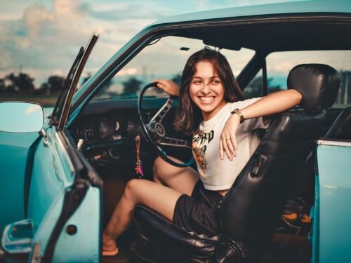 A cheerful young woman sitting in a blue vintage convertible car outdoors during the day.