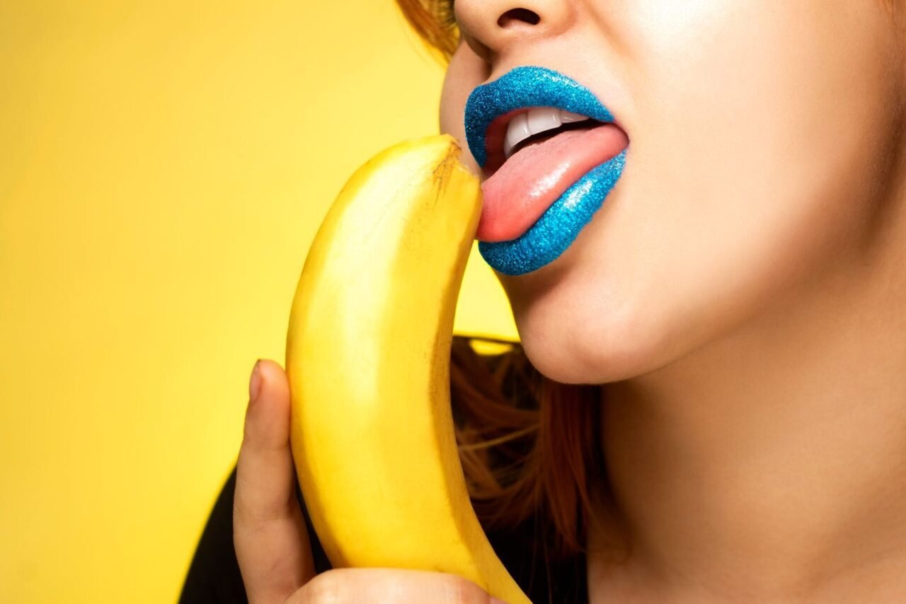 Close-up of vibrant blue glitter lips near a banana against a yellow background.