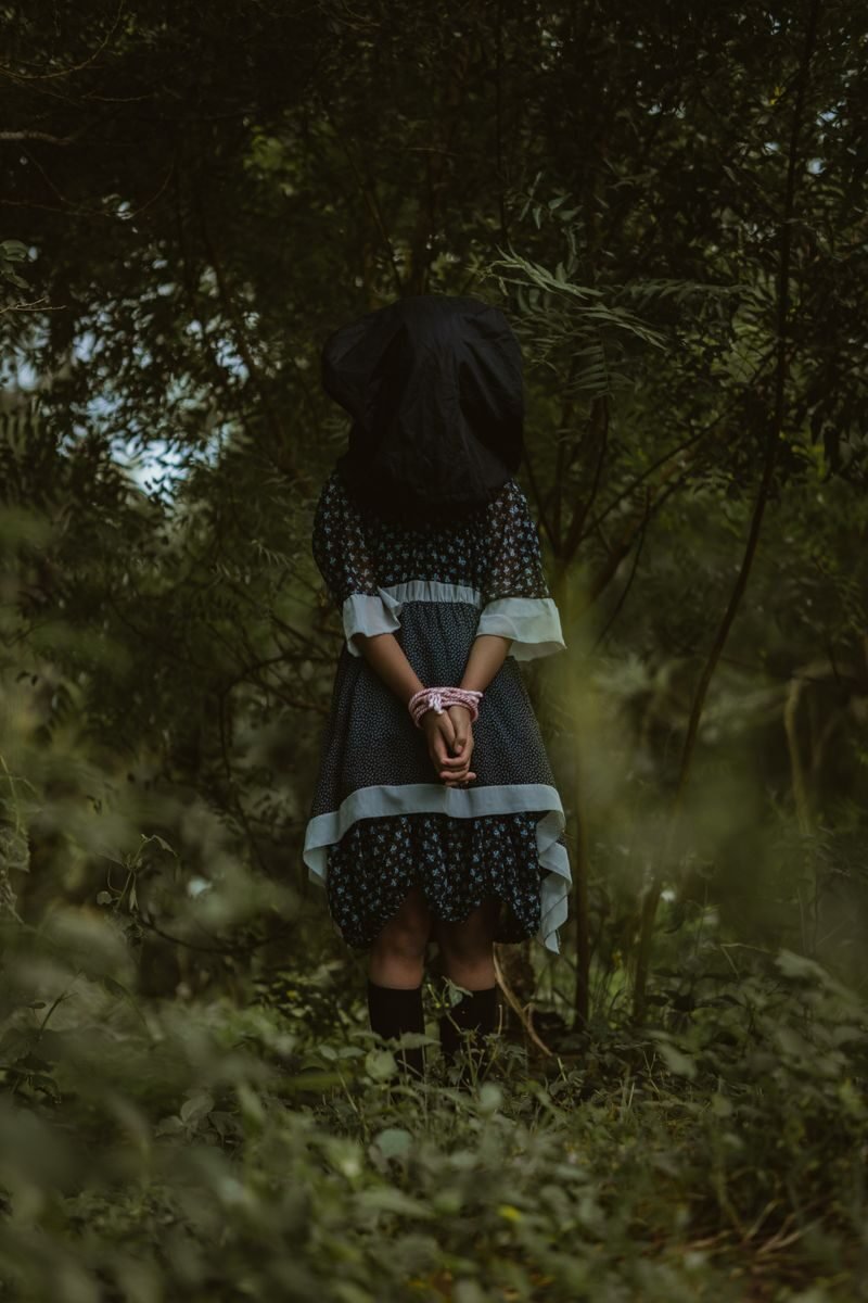 A woman in a dark dress poses mysteriously in a dense forest, hands tied.