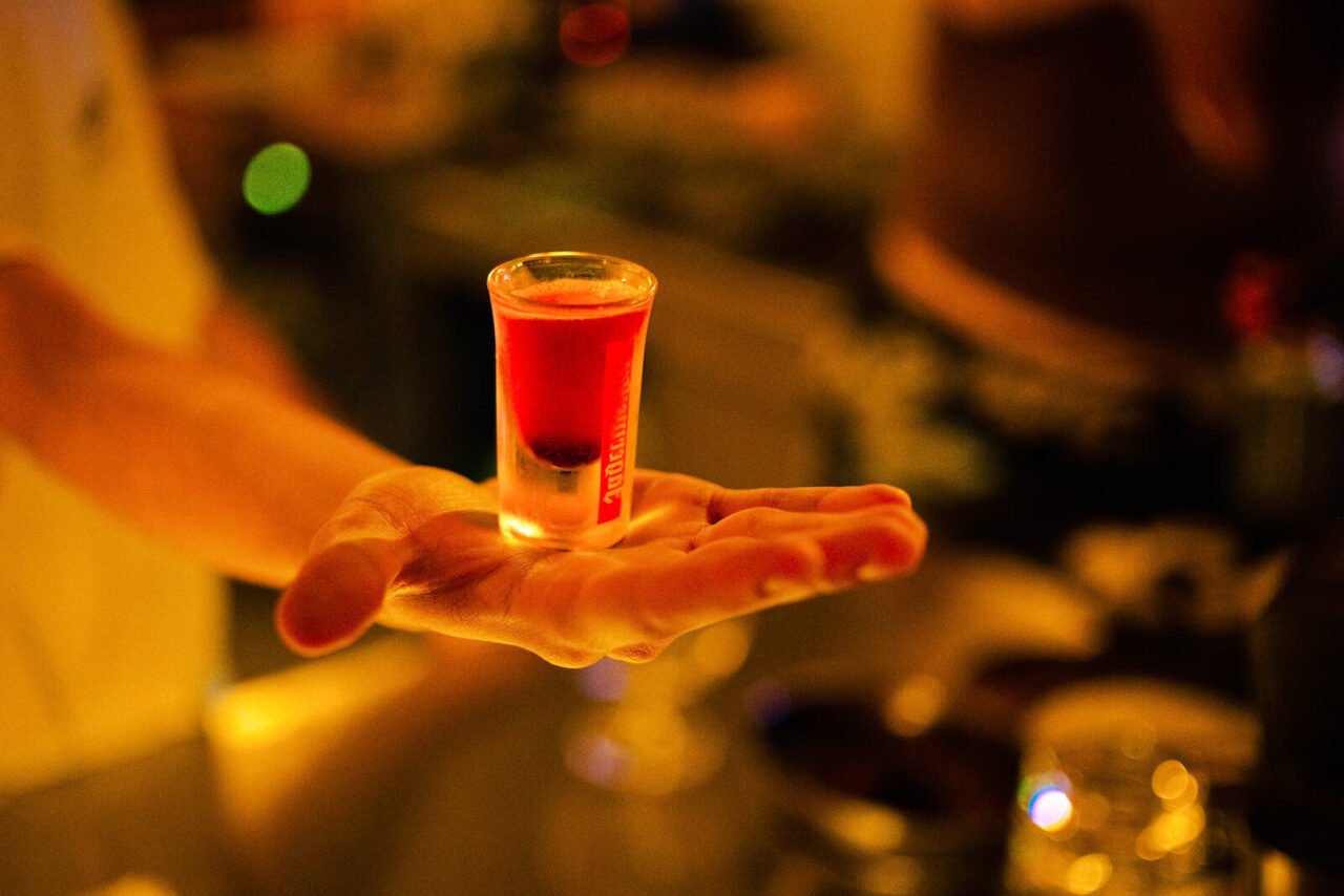 A vibrant red cocktail shot gracefully balanced on a person's hand in a dimly lit bar setting.