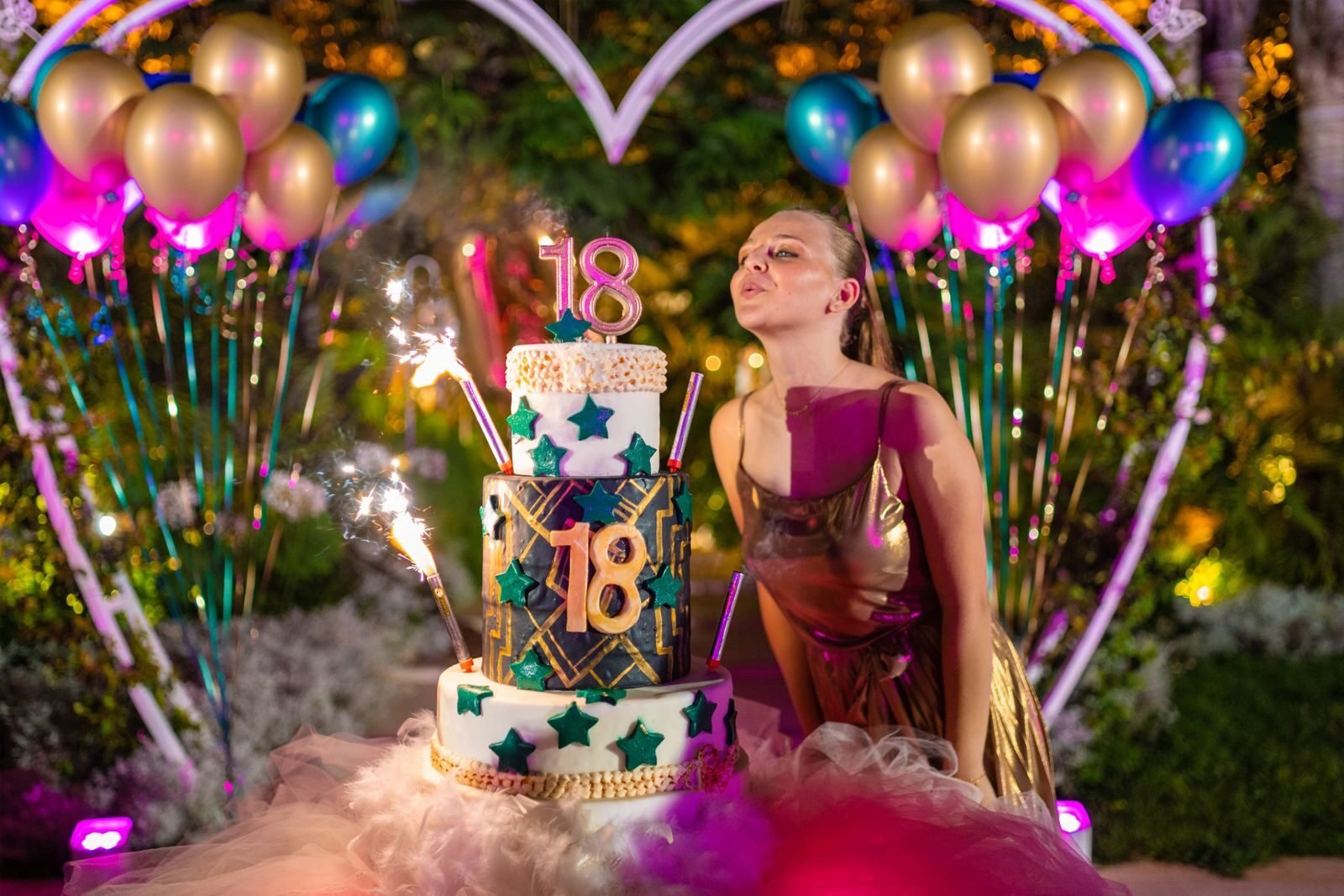 Content young female in elegant dress blowing shiny candles on birthday cake while standing with pouting lips near decorative heart and colorful balloons during party