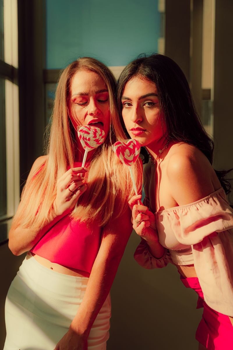 Two young women enjoy lollipops in warm indoor light, embodying playful fashion and friendship.