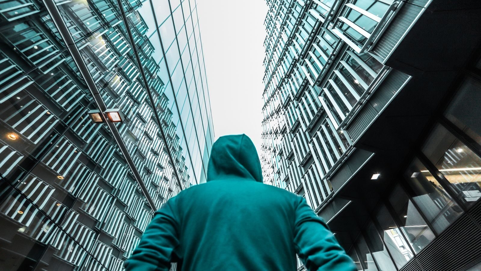A person in a hoodie looks up at sleek, reflective skyscrapers in a cityscape.