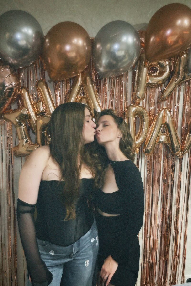 Two women share a kiss at a birthday party in front of festive balloons.