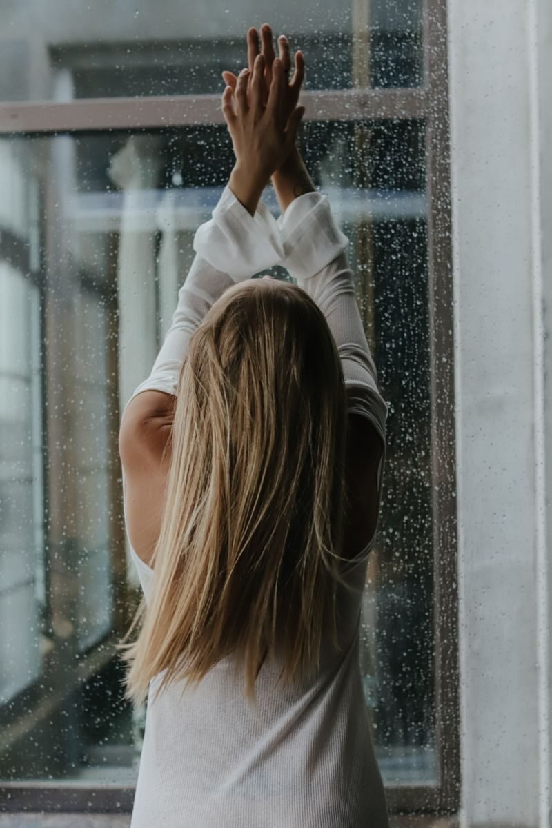 A woman with long blonde hair stretching by a window on a rainy day, evoking a calm and introspective mood.