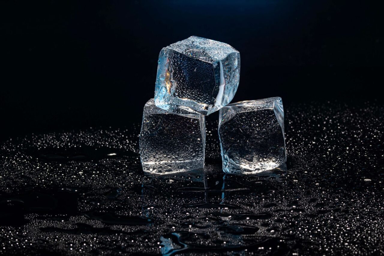Three clear ice cubes stacked in a dark setting with water droplets for a cooling effect.