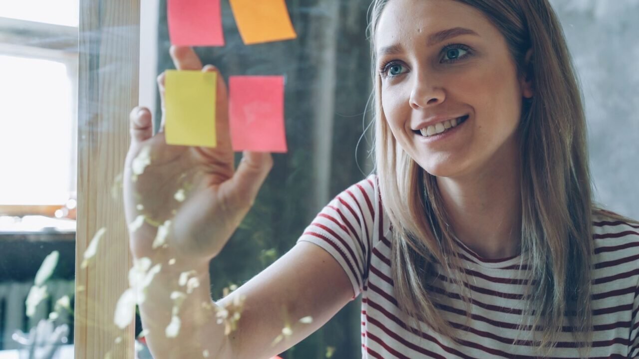 Smiling woman in striped shirt arranging colorful sticky notes on glass.