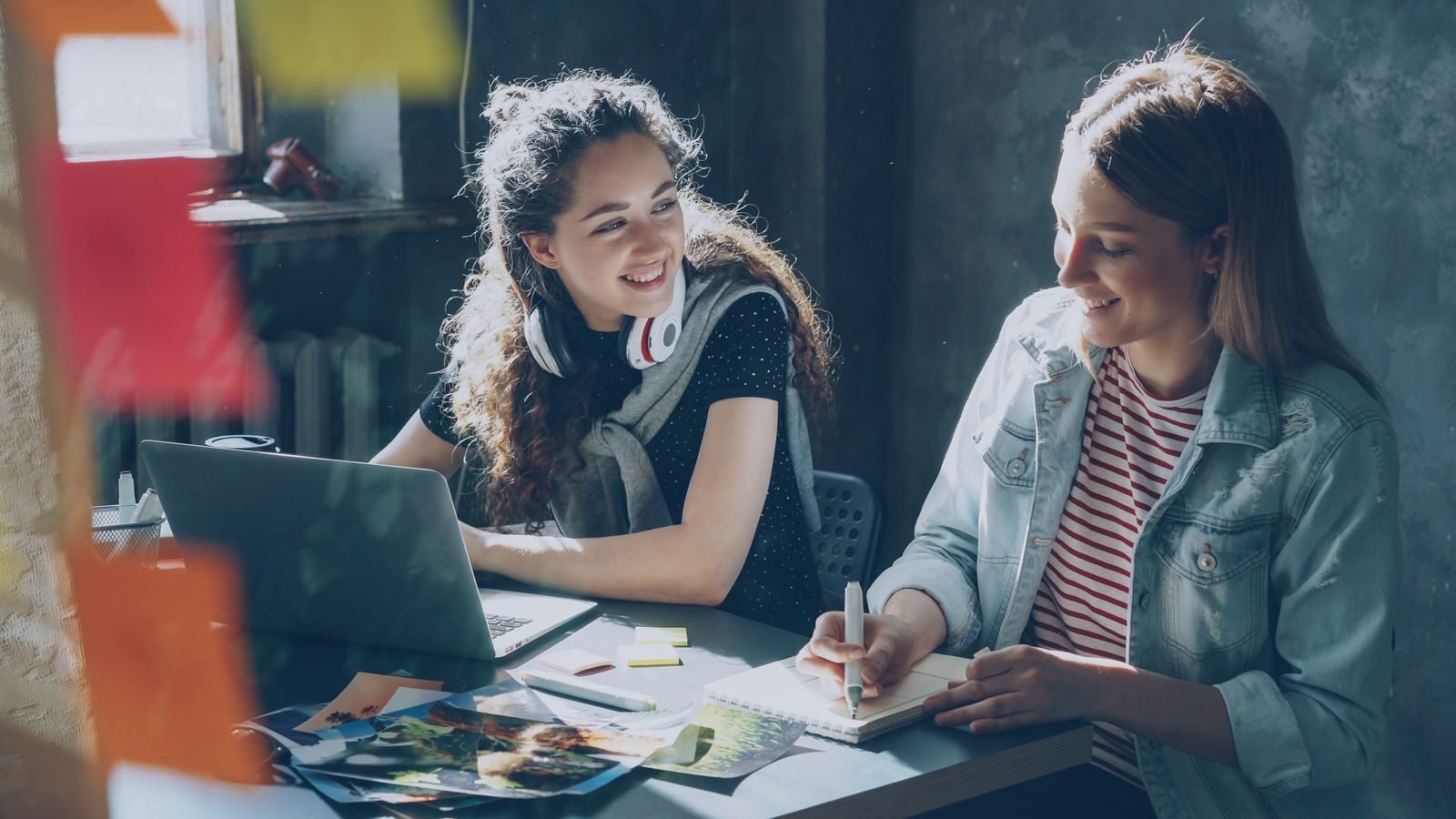 Two young women smiling and working together in a creative indoor setting with a laptop.