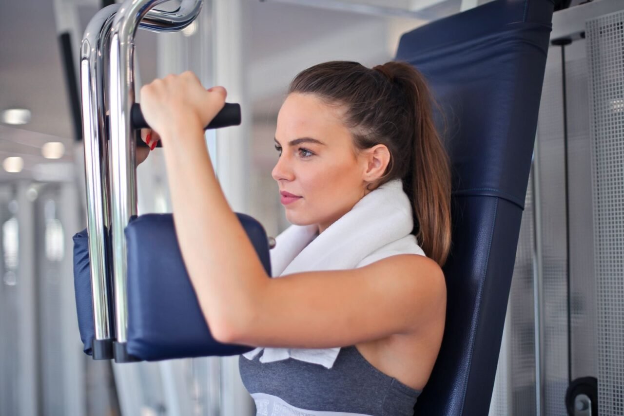 Slender focused woman in sportswear with towel on neck performing exercise on weight machine in light contemporary gym