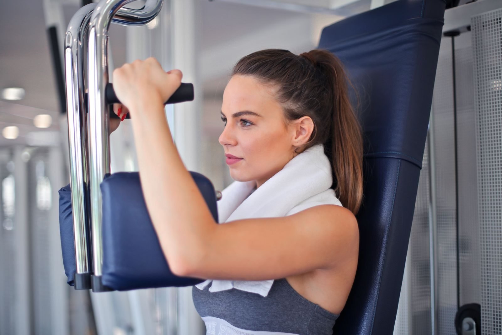 Slender focused woman in sportswear with towel on neck performing exercise on weight machine in light contemporary gym