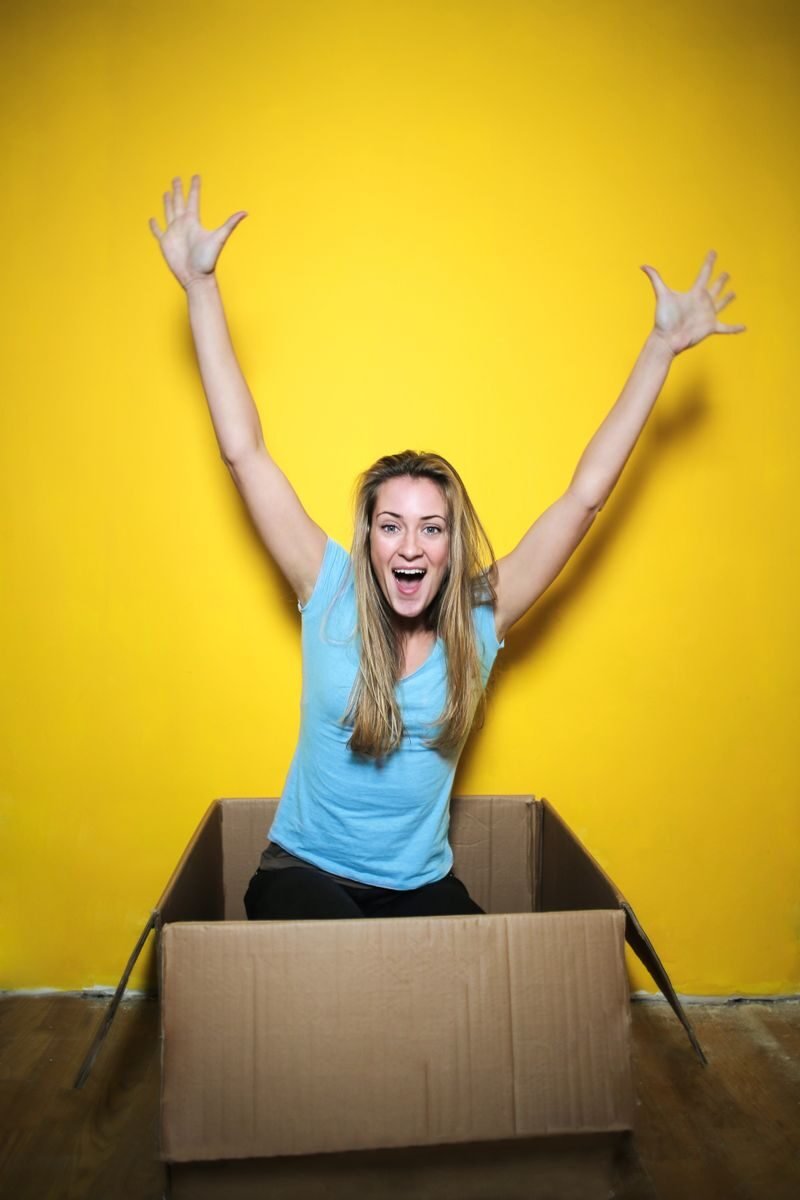 Young woman with natural hair celebrating in a cardboard box against a yellow background.