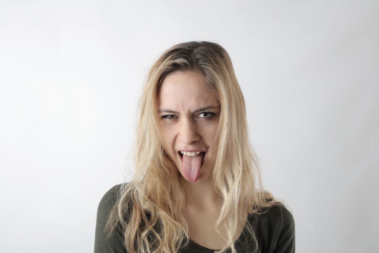 A playful woman with blond hair makes a funny face, sticking her tongue out on a white background.