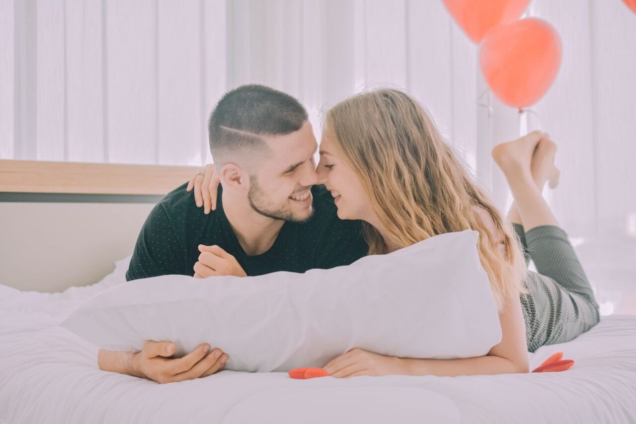 Affectionate couple lying in bed, surrounded by romantic red balloons, creating a warm and intimate scene.