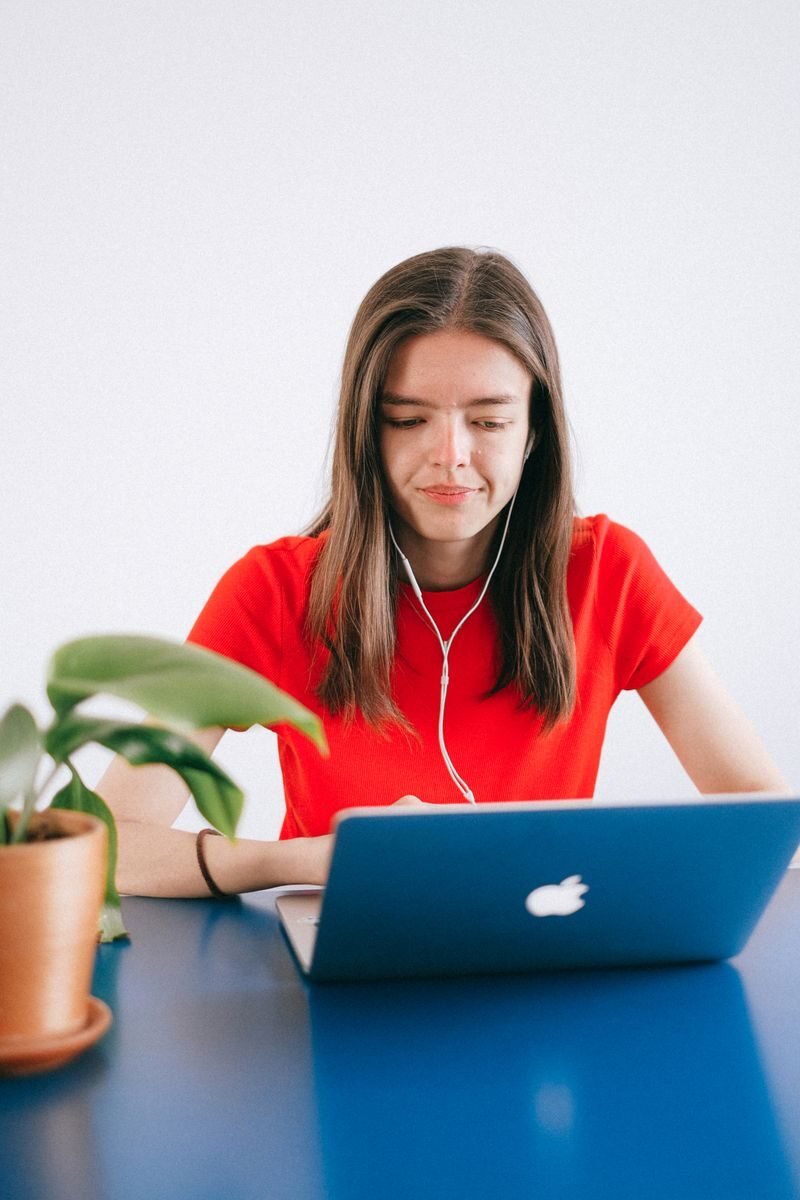 Focused young woman with headphones working on a laptop at a blue table, surrounded by plants.