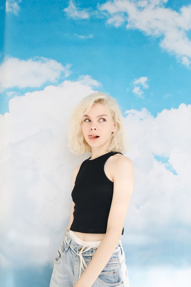 Playful blonde woman in a black crop top and jeans posing against a bright blue sky background.