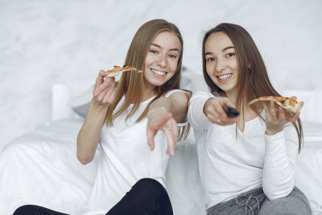 Two young women smiling, enjoying pizza, and pointing a remote control in a cozy indoor setting.