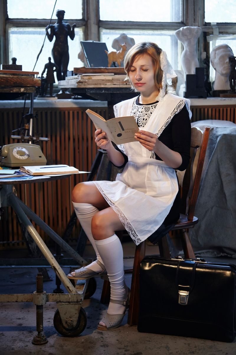 Full body of concentrated female wearing soviet school uniform reading book while sitting at table with retro telephone in USSR styled room