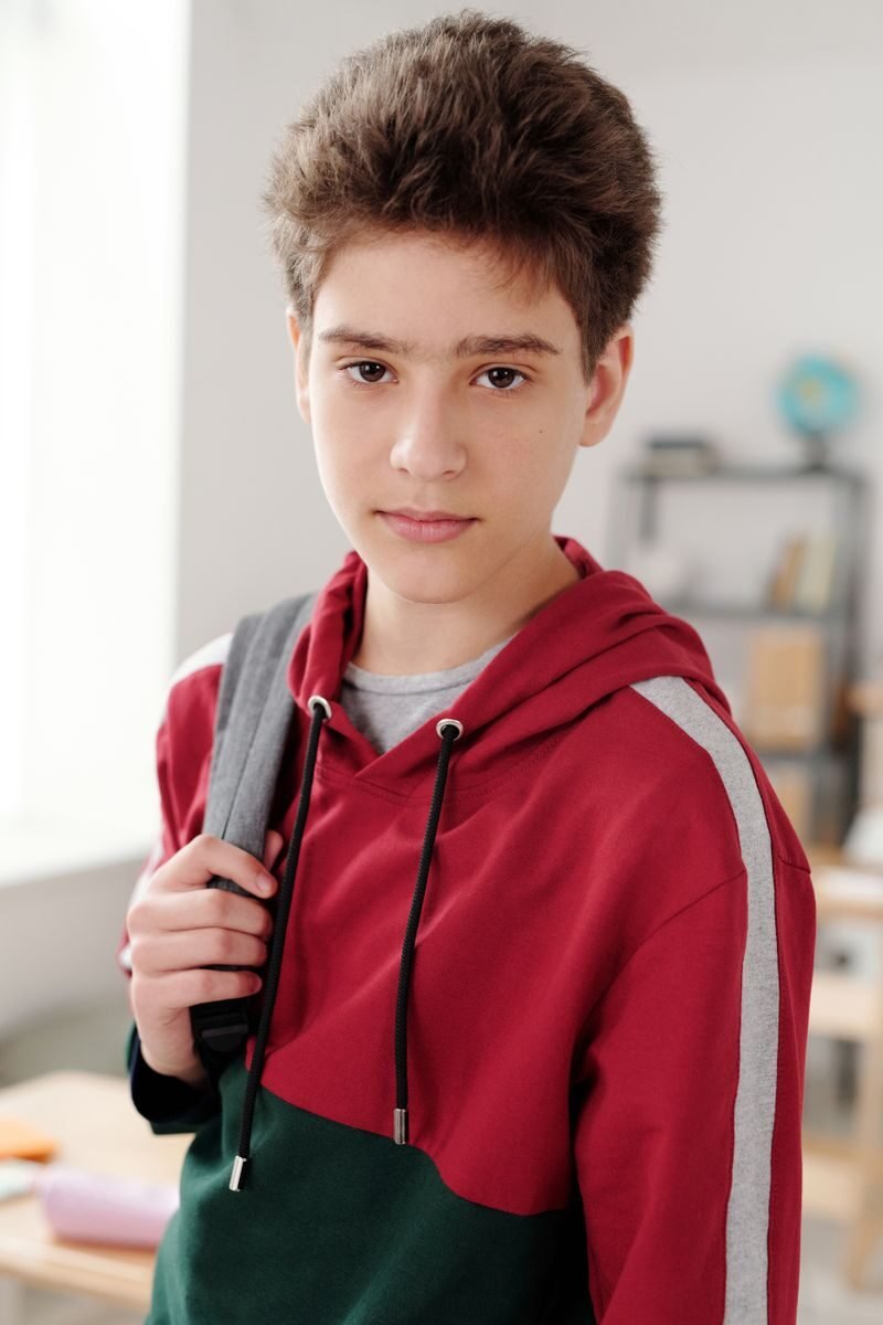 Teen boy in a colorful hoodie holding a backpack, standing in a classroom.