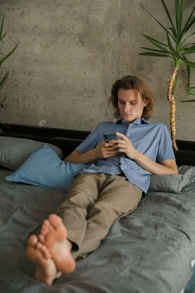 Relaxed barefoot male in casual wear lying on comfortable bed while messaging on mobile phone and looking at screen