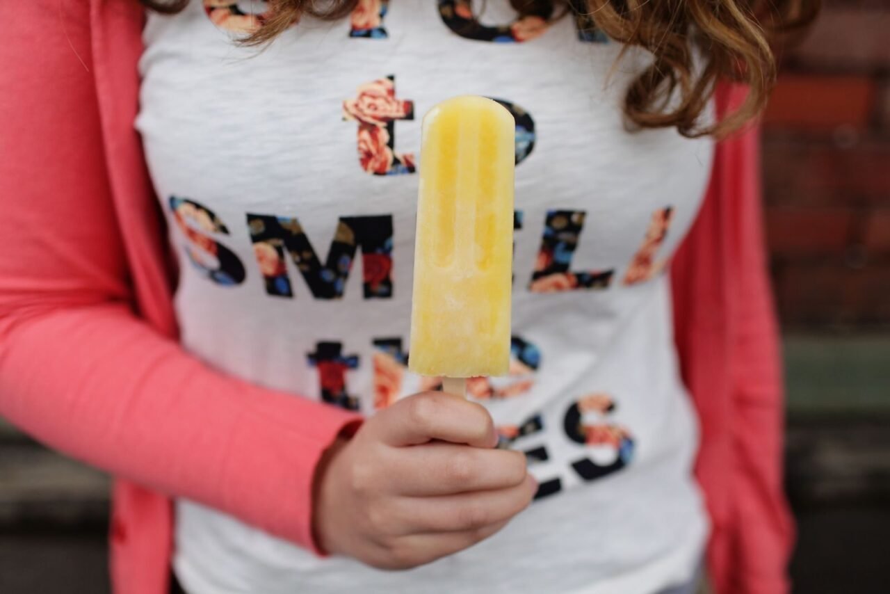Close-up of a child holding a bright yellow popsicle with a colorful shirt and jacket.