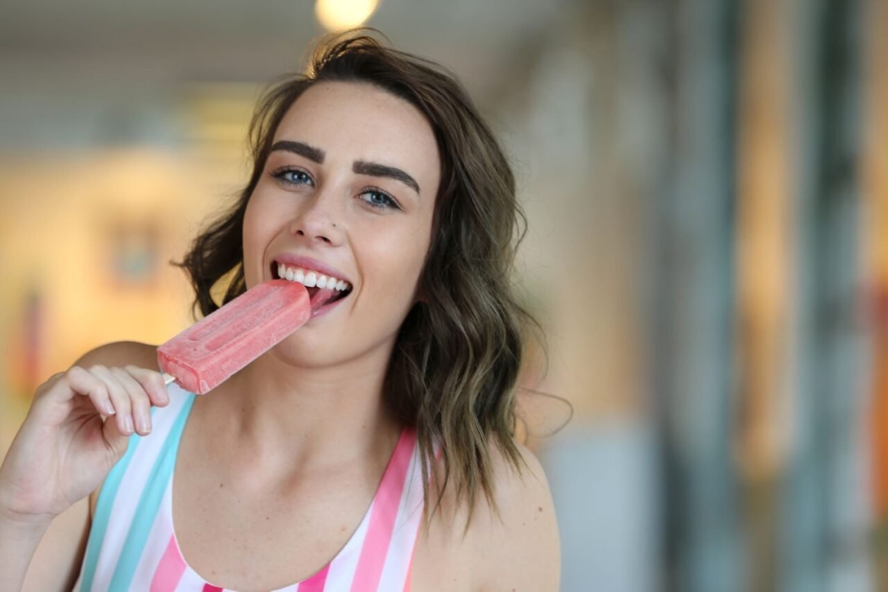Smiling woman savoring a pink popsicle inside, capturing a moment of joy and refreshment.