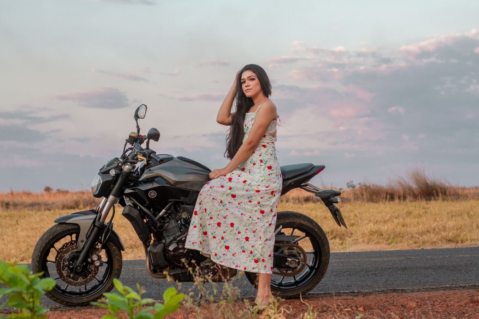 A woman in a floral dress sits on a motorcycle parked by the roadside during sunset.