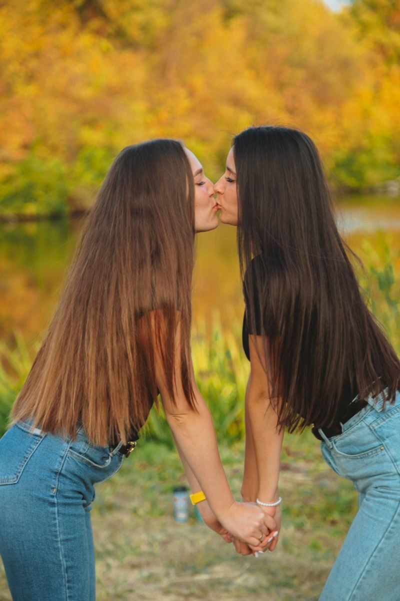 Side view of young women in casual clothes holding hands and kissing each other on river bank