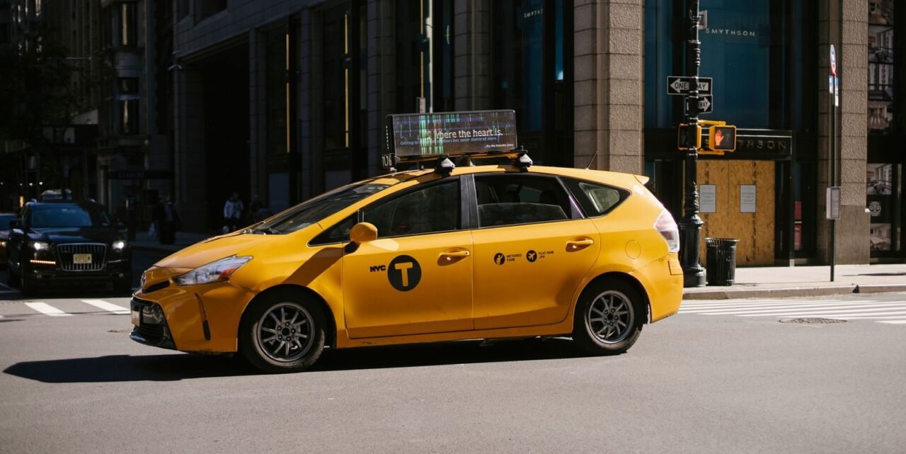 Side view of yellow cab with logo on door riding along modern building and crossroad on asphalt road in street