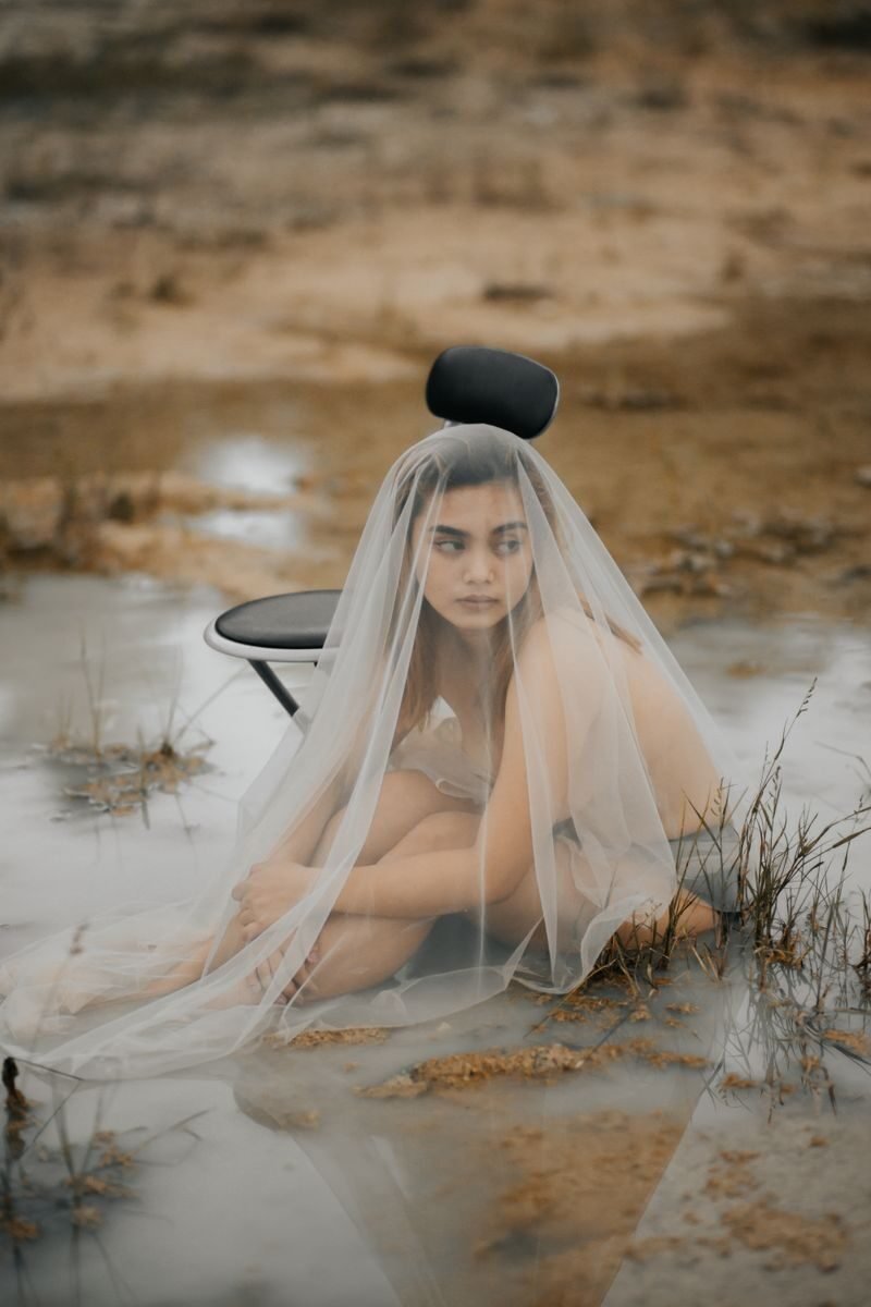 Young pondering ethnic female in transparent textile sitting in pond near chair while looking away
